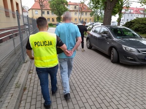 policjant w kamizelce odblaskowej z napisem policja prowadzi mężczyznę skutego kajdankami