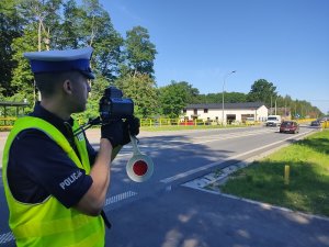 przy drodze stoi umundurowany policjant ruchu drogowego. Ma na sobie kamizelkę odblaskową. W rękach trzyma tarczę do zatrzymywania pojazdów i ręczny miernik prędkości, który wycelowany jest w jadący drogą samochód osobowy