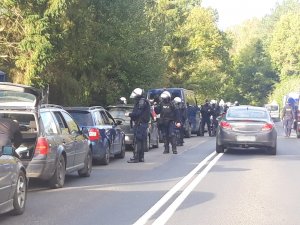 policjanci wyposażeni są w zestaw przeciwuderzeniowy, który składa się z kamizelki przeciwuderzeniowej z napisem POLICJA wraz z ochraniaczami barków i ramion, ochraniacze nóg chroniące kolano, goleń a także podbicie stopy, ochraniacze przedramienia. Każdy z policjantów posiada również kask przeciwuderzeniowy z przyłbicą ochraniajacą twarz, osłonę twarzy oraz pałkę szturmową typu PS. Stoją przy cywilnych samochodach osobowych, ustawionych w rzędzie wzdłuż ulicy