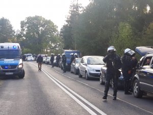 umundurowani policjanci wyposażeni są w zestaw przeciwuderzeniowy, który składa się z kamizelki przeciwuderzeniowej z napisem POLICJA wraz z ochraniaczami barków i ramion, ochraniacze nóg chroniące kolano, goleń a także podbicie stopy, ochraniacze przedramienia. Każdy z policjantów posiada również kask przeciwuderzeniowy z przyłbicą ochraniajacą twarz, osłonę twarzy oraz pałkę szturmową typu PS. Stoją przy cywilnych samochodach osobowych ustawionych wzdłuż ulicy. W jednym samochodzie szyba jest uchylona i widać głowę pasażera. Po drugiej stronie ulicy stoi policyjny oznakowany radiowóz z uruchomionymi sygnałami świetlnymi. Wzdłuż drogi idzie mężczyzna w cywilnym ubraniu