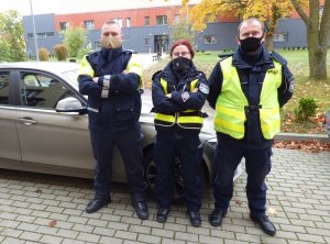 Troje umundurowanych policjantów ruchu drogowego, w tym jedna kobieta, stoi w rzędzie przed nieoznakowanym radiowozem. Policjanci mają na twarzach maseczki ochronne