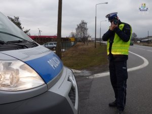 Umundurowany policjant ruchu drogowego stoi przy drodze obok radiowozu. Dokonuje pomiaru prędkości ręcznym miernikiem prędkości