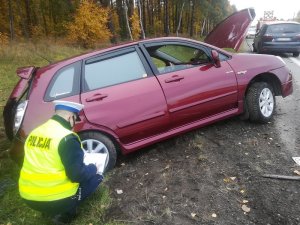 umundurowany policjant ruchu drogowego kuca przy znajdującym się w rowie, po lewej stronie drogi k6, uszkodzonym bordowym osobowym suzuki. Policjant wypełnia dokument