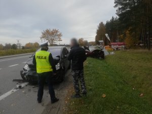 na poboczu po prawej stronie drogi k6 stoi ciemnoszary ford s-max. Przedni zderzak tego samochodu jest połamany, reflektory uszkodzone. Za fordem w rowie znajduje się uszkodzone bordowe osobowe suzuki. Maska suzuki jest otwarta. Przed fordem stoi tyłem umundurowany policjant ruchu drogowego oraz mężczyzna w ciemnej kurtce i spodniach ze wzorem moro