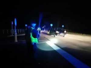 wieczorowa pora. Na ulicy za oznakowanym przejściem dla pieszych stoi porsche cayenne. Na pierwszym planie, po lewej stronie drogi stoi umundurowany policjant ruchu drogowego. Przy porsche widoczne trzy osoby w kamizelkach odblaskowych