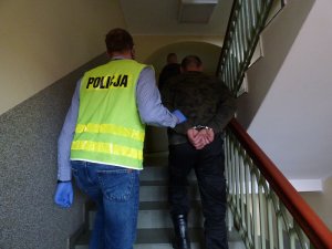 policjant w cywilnym ubraniu i odblaskowej kamizelce z napisem policja prowadzi po schodach mężczyznę ze skutymi kajdankami rękoma