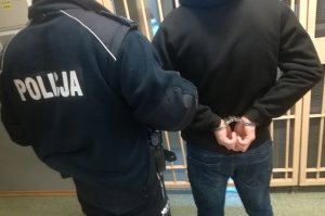 korytarz pomieszczenia dla osób zatrzymanych. W tle kraty. Na pierwszym planie umundurowany policjant trzyma za przedramię skutego kajdankami mężczyznę w czarnej bluzie z kapturem. Obydwaj ustawieni tyłem do obiektywu