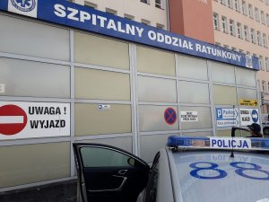 przed szpitalnym oddziałem ratunkowym widoczny jest fragment oznakowanego radiowozu policyjnego. Drzwi radiowozu od strony kierowcy są otwarte. Przy drzwiach od strony pasażera stoi policjantka, która spogląda w kierunku wiaty SOR