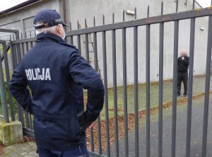 przed metalowym ogrodzeniem stoi tyłem do obiektywu umundurowany policjant z maseczką ochronną na twarzy. Za ogrodzeniem obok budynku o szarej elewacji stoi zwrócony przodem do policjanta mężczyzna w czarnej bluzie i ciemnych spodniach.