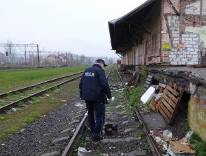 zwrócony tyłem do obiektywu umundurowany policjant idzie wzdłuż torów kolejowych w kierunku starej, zniszczonej wyładowni. Policjant spogląda na dół na zasłonięte fragmentami drewnianych palet otwory w betonowej części wyładowni. Okolica zaśmiecona jest kartonami, folią i plastikiem