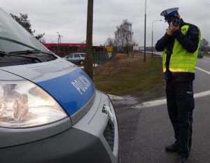 na poboczu drogi stoi przodem do obiektywu umundurowany policjant ruchu drogowego w kamizelce odblaskowej. Policjant mierzy prędkość za pomocą ręcznego miernika prędkości. Obok niego stoi zaparkowany oznakowany radiowóz fiat ducato