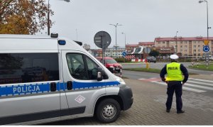 przed przejściem dla pieszych stoi skierowany tyłem do obiektywu umundurowany policjant ruchu drogowego w kamizelce odblaskowej. Obok niego stoi oznakowany radiowóz. W tle widać rondo, budynki mieszkalne i jadące ulicą samochody