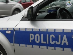 zdjęcie przedstawia fragment oznakowanego radiowozu z widocznym napisem bocznym policja. Na desce rozdzielczej radiowozu leży czapka policjanta ruchu drogowego. W tle widoczne przejeżdżające ulicą samochody