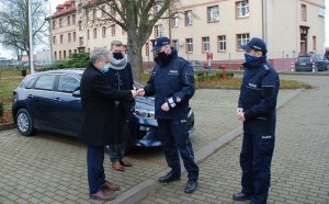 na parkingu od wewnętrznej strony terenu Komendy Powiatowej Policji w Lęborku Burmistrz Miasta Lęborka - mężczyzna w czarnym płaszczu, wręcza kluczyki Komendantowi Powiatowemu Policji w Lęborku ubranemu w mundur służbowy. Obok komendanta, po jego lewej stronie stoi umundurowany policjant w stopniu komisarza. Nieco w tyle, pomiędzy burmistrzem i komendantem stoi mężczyzna w czarnej kurtce. Wszyscy mają maseczki ochronne na twarzach. Mężczyźni stoją przed nieoznakowanym radiowozem kia ceed granatowego koloru. W tle widoczny budynek komendy policji