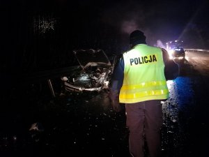 na dworze jest ciemno. Na ulicy na mokrej nawierzchni stoi skierowany tyłem do obiektywu umundurowany policjant. Policjant stoi przed samochodem osobowym srebrnego koloru. Maska samochodu jest otwarta. Przód auta zgnieciony