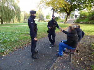 w miejskim parku dwaj umundurowani policjanci z kominami zasłaniającymi usta i nos stoją przed ławką na której siedzą trzy osoby, dwóch mężczyzn i kobieta. Mężczyzna z maseczką ochronną na twarzy, siedzący po lewej stronie ławki, podaje policjantowi dowód osobisty