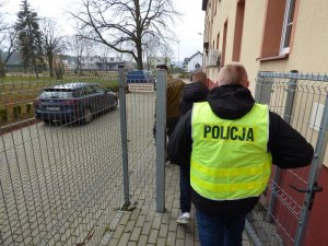 policjanci wyprowadzają z budynku komendy zatrzymanego mężczyznę. Wszyscy są zwróceni tyłem do obiektywu. Na pierwszym planie znajduje się nieumundurowany policjant w kamizelce odblaskowej z napisem policja. Przed nim idzie zatrzymany mężczyzna w czarnej kurtce z kapturem. Jako pierwszy idzie nieumundurowany policjant. Wszyscy kierują się w stronę nieoznakowanego radiowozu znajdującego się na wewnętrznym parkingu komendy