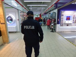 wnętrze sklepu kaufland. Skierowany tyłem do obiektywu umundurowany policjant idzie w kierunku wyjścia z kas sklepowych. Po obu jego stronach znajdują się punkty handlowe. W tle, przy kasach, znajdują się klienci w maseczkach ochronnych