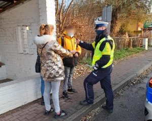 przed przystankiem autobusowym z białej cegły stoi skierowany lewym bokiem do obiektywu umundurowany policjant ruchu drogowego z zasłoniętymi ustami i nosem. Policjant w lewej dłoni trzyma opaskę odblaskową. Przed nim na chodniku stoją trzy osoby: młoda kobieta w złotej kurtce, z blond włosami i w maseczce ochronnej i kilkunastoletni chłopak w pomarańczowo - czarnej kurtce. Trzecia osoba jest zasłonięte przez kobietę. Policjant wyciąga rękę z opaską odblaskową w kierunku kobiety