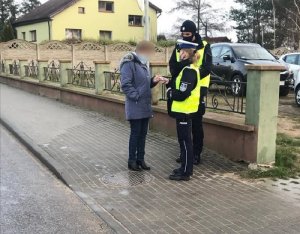 na chodniku przy murowanym niskim ogrodzeniu z metalowymi elementami, stoją umundurowani policjant i policjantka ruchu drogowego oraz kobieta w szarej zimowej kurtce. Wszyscy mają zasłonięte usta i nos. Policjantka przekazuje kobiecie element odblaskowy. W tle parking z osobowymi samochodami i budynek mieszkalny z żółtą elewacją