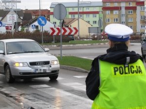 Ulica na terenie miasta. Na trawie znajduje się topniejący śnieg. Z ronda zjeżdża osobowe audi w srebrnym kolorze. Jest przed przejściem dla pieszych. Na chodniku przed przejściem dla pieszych stoi umundurowana policjantka ruchu drogowego w kamizelce odblaskowej.