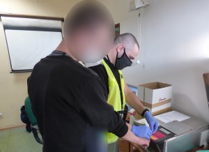 pomieszczenie techników kryminalistyki. Nieumundurowany policjant w kamizelce odblaskowej, rękawiczkach ochronnych i maseczce ochronnej na twarzy zdejmuje odciski palców od zatrzymanego mężczyzny w czarnej bluzie. Głowa mężczyzny jest zasłonięta filtrem zamglenia obrazu