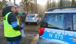 przy radiowozie stoi nieumundurowany policjant w kamizelce odblaskowej. Na rękawie ma opaskę z napisem negocjator. Policjant trzyma w dłoni megafon. Obok niego stoi umundurowany żołnierz