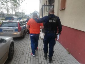 umundurowany policjant prowadzi zatrzymanego mężczyznę. Trzyma lewą rękę na karku mężczyzny. Kierują się w stronę radiowozu