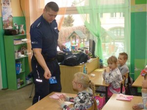 klasa szkolna. przy ławkach siedzą dzieci. Przed nimi stoi umundurowany policjant. Policjant podaje dziewczynce ulotkę z logo policji