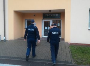 skierowani tyłem do obiektywu dwaj umundurowani policjanci wchodzą po schodach prowadzących do zakładu fryzjerskiego. Przed nimi idą dwie pracownice sanepidu