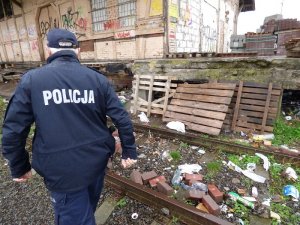 umundurowany policjant idzie wzdłuż starej nieczynnej rampy kolejowej znajdującej się przy torach. Teren jest zaśmiecony, przy rampie ustawione są drewniane palety, zasłaniające otwory w betonowych blokach