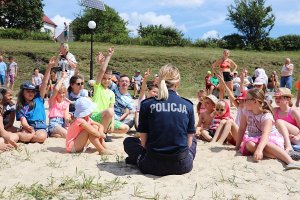 na plaży dzieci wysłuchują prelekcji umundurowanej policjantki, która siedzi z nimi na piasku. Większość dzieci zgłasza się do odpowiedzi