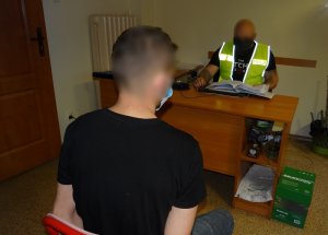 Pokój przesłuchań. Na krześle przed biurkiem siedzi zatrzymany mężczyzna, przed nim, za biurkiem siedzi policjant w kamizelce odblaskowej z napisem policja