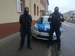 przed budynkiem policji, przed oznakowanym radiowozem stoją dwaj umundurowani policjanci patrolówki. Usta i nos mają zasłonięte kominami ochronnymi