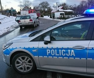 na pierwszym planie, wszerz drogi stoi oznakowany radiowóz. Za nim przy lewej stronie drogi znajduje się szary vw golf z otwartą maską i otwartymi drzwiami od strony kierowcy. Po prawej stronie drogi, poza jezdnią znajduje się biały bus. Obok niego wóz straży pożarnej