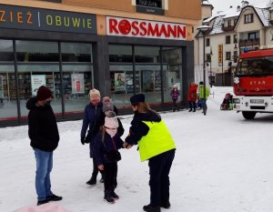 umundurowana policjantka nakłada na rękę dziewczynki opaskę odblaskową. Za dziewczynką stoi młodsze dziecko i dwie osoby dorosłe.