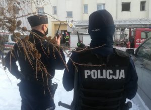 przed blokiem w którym doszło do pożaru stoją dwaj umundurowani policjanci. Jeden z nich rozmawia przez radiostację. Przed nimi stoi wóz strażacki