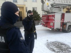 przed budynkiem, w który doszło do pożaru stoją dwaj umundurowani policjanci. Jeden z nich rozmawia przez telefon. Przed budynkiem stoi wóz strażacki