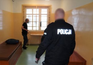 pomieszczenie dla osób zatrzymanych. Przy oknie stoi skierowany bokiem do obiektywu zatrzymany mężczyzna. Przed nim, przy drzwiach do celi stoi umundurowany policjant