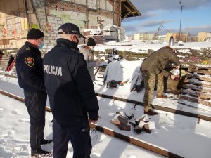 teren starej nieczynnej wyładowni przy dworcu PKP. Przy torach kolejowych, przed wyładownią stoją policjant, funkcjonariusz straży miejskiej i dwoje pracowników miejskiego ośrodka pomocy społecznej: kobieta i mężczyzna. Mężczyzna nalewa do kubka napój i podaje osobie schowanej pod betonową rampą