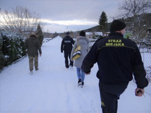 teren ogródków działkowych. Ścieżką, wzdłuż działek idą policjant, funkcjonariusz straży miejskiej i dwoje pracowników socjalnych: kobieta i mężczyzna