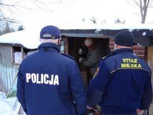 teren ogródków działkowych. Przed altanką stoją policjant oraz funkcjonariusz straży miejskiej. przed drzwiami altanki stoi pracownik socjalny, który nalewa do kubka napój