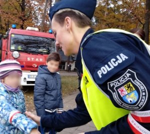 umundurowana policjantka wręcza dziewczynce opaskę w biało-czerwonych barwach. Obok stoi chłopiec. W tle znajduje się wóz straży pożarnej