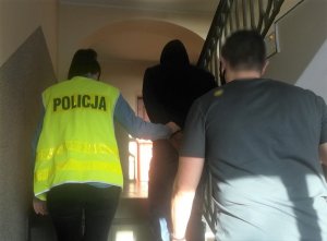 Policjantka w kamizelce odblaskowej i policjant w ubraniu cywilnym prowadzą po schodach zatrzymanego mężczyznę