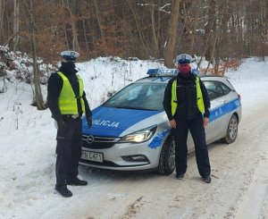 przed oznakowanym radiowozem na ośnieżonej drodze przy lesie stoją umundurowani policjant i policjantka w kamizelkach odblaskowych