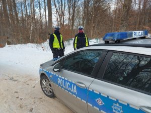 przed oznakowanym radiowozem na ośnieżonej drodze przy lesie stoją umundurowani policjant i policjantka w kamizelkach odblaskowych