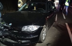 pora wieczorowa. Na drodze stoi czarne osobowe bmw z rozbitym przodem. Przy samochodzie znajdują się funkcjonariusze straży pożarnej i policjant