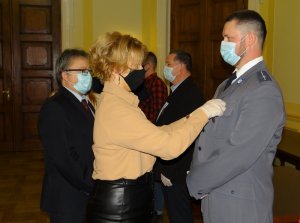 aula starostwa powiatowego w Lęborku. Przed policjantem w mundurze galowym stoi kobieta, która przypina mu do klapy marynarki odznakę. Obok niej stoi wicestarosta lęborski