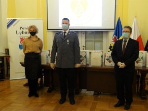 aula starostwa. Przed stołem, na tle flagi Polski i powiatu lęborskiego stoi policjant w mundurze galowym. Po jego obu stronach stoją kobieta i mężczyzna. Wszyscy mają maseczki ochronne na twarzach