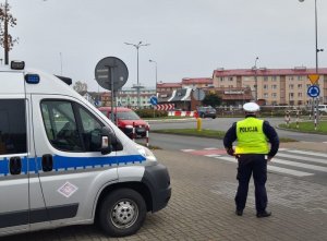 na chodniku przed przejściem dla pieszych, obok oznakowanego radiowozu stoi umundurowany policjant ruchu drogowego, który obserwuje ruch uliczny